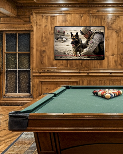 A cozy wooden-paneled room with a pool table in the center with balls racked up for a game. Above the table on the back wall hangs a framed photo of a military soldier beside a German Shepherd dog in a desert setting.